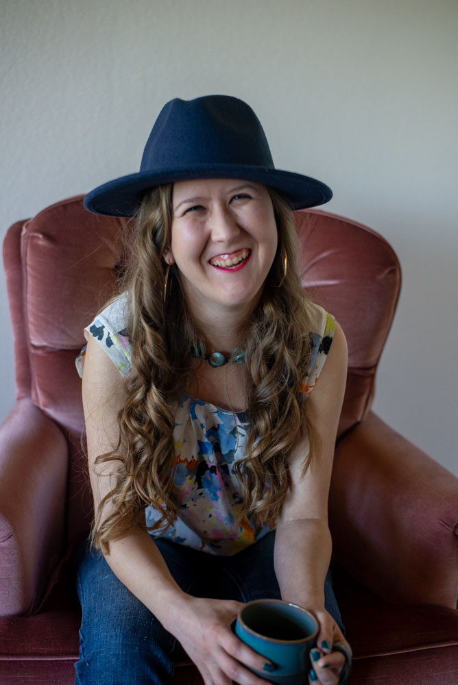 Owner Christina Damron sitting down with a cup of coffee in a velvet pink chair wearing a teal hat.