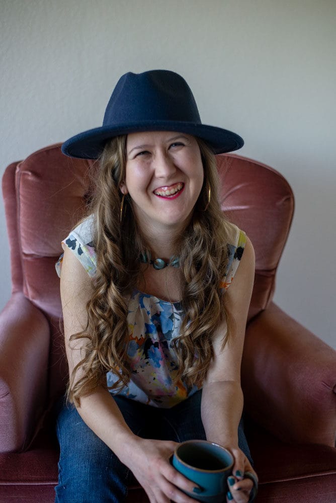 Owner Christina Damron sitting down with a cup of coffee in a velvet pink chair wearing a teal hat.